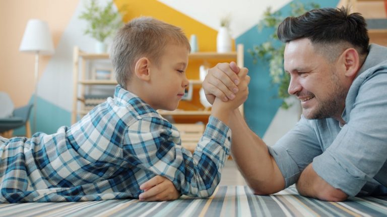 Adorable kid is wrestling with joyful father having fun winning indoors in apartment lying on carpet on floor. Family activity and healthy lifestyle concept.